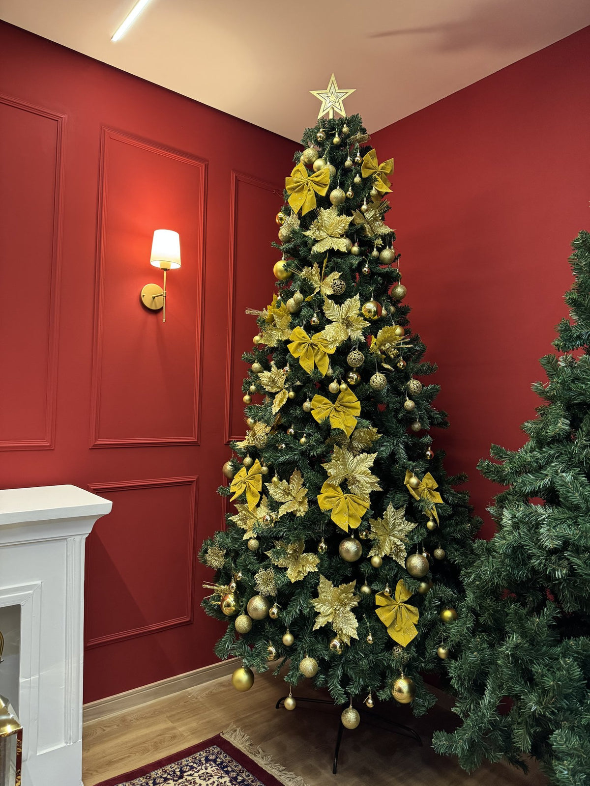 Pinheiro canadense tradicional de 2,70 m decorada com flores e laços amarelos, bolas douradas e estrela, em canto de sala com parede vermelha