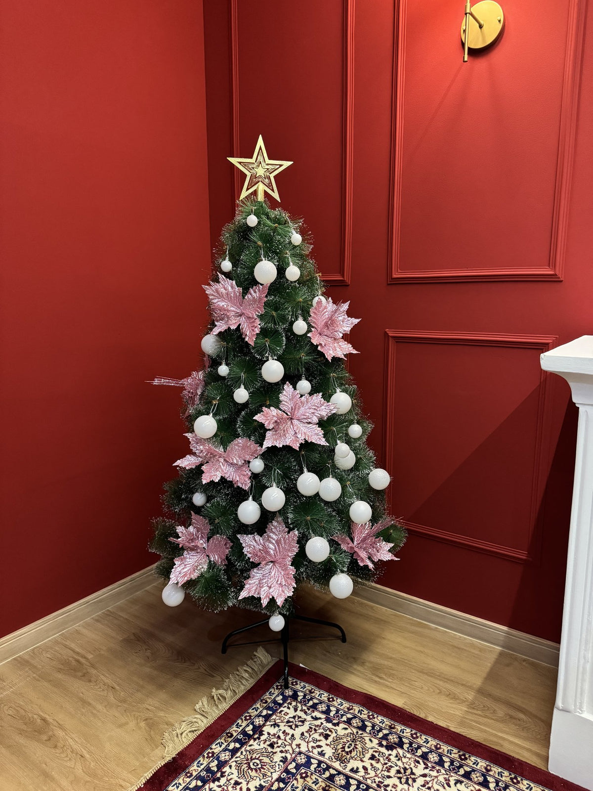 Árvore pompom com pontas nevadas de 1,50 m decorada com bolas brancas, flores rosa e estrela dourada, em canto de sala com parede vermelha