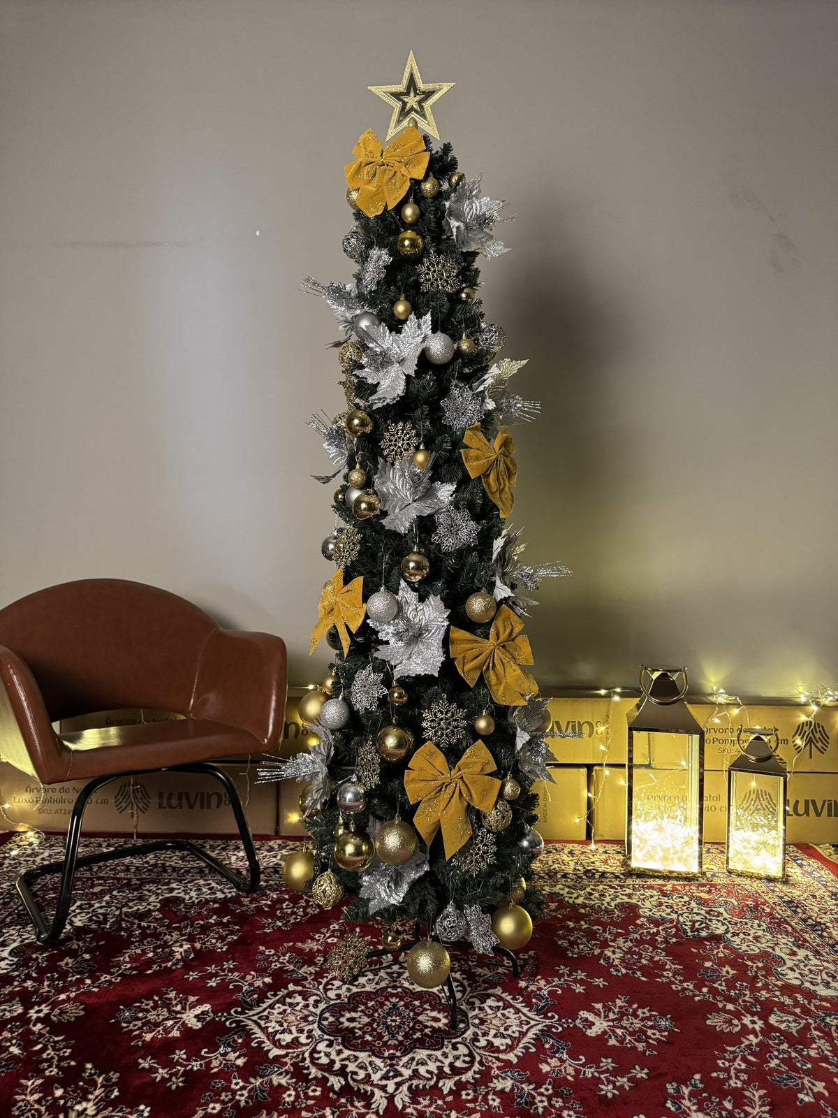 Árvore slim de 1,80 m decorada com laços e bolas douradas, flores prateadas e estrela, ao lado de uma poltrona de couro