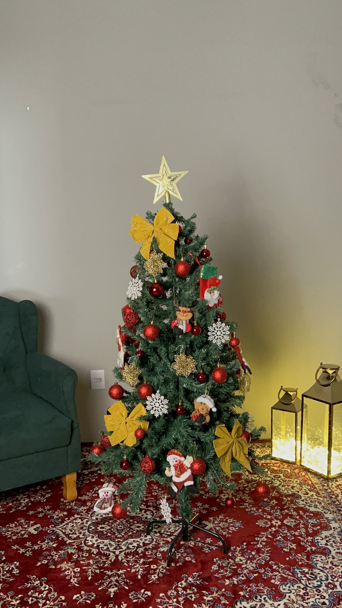 Pinheiro canadense tradicional de 1,50 m decorada com laços dourados, bolas vermelhas e estrela, ao lado de poltrona verde e lanternas