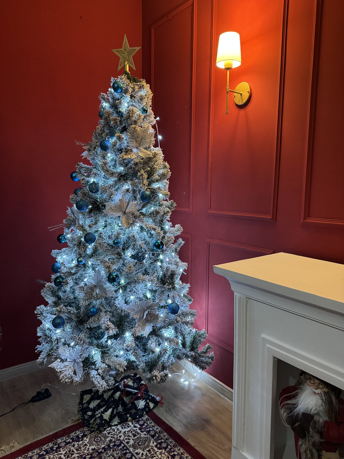 Árvore nevada de 1,80 m decorada com bolas azuis, laços dourados e luzes, em sala com parede vermelha