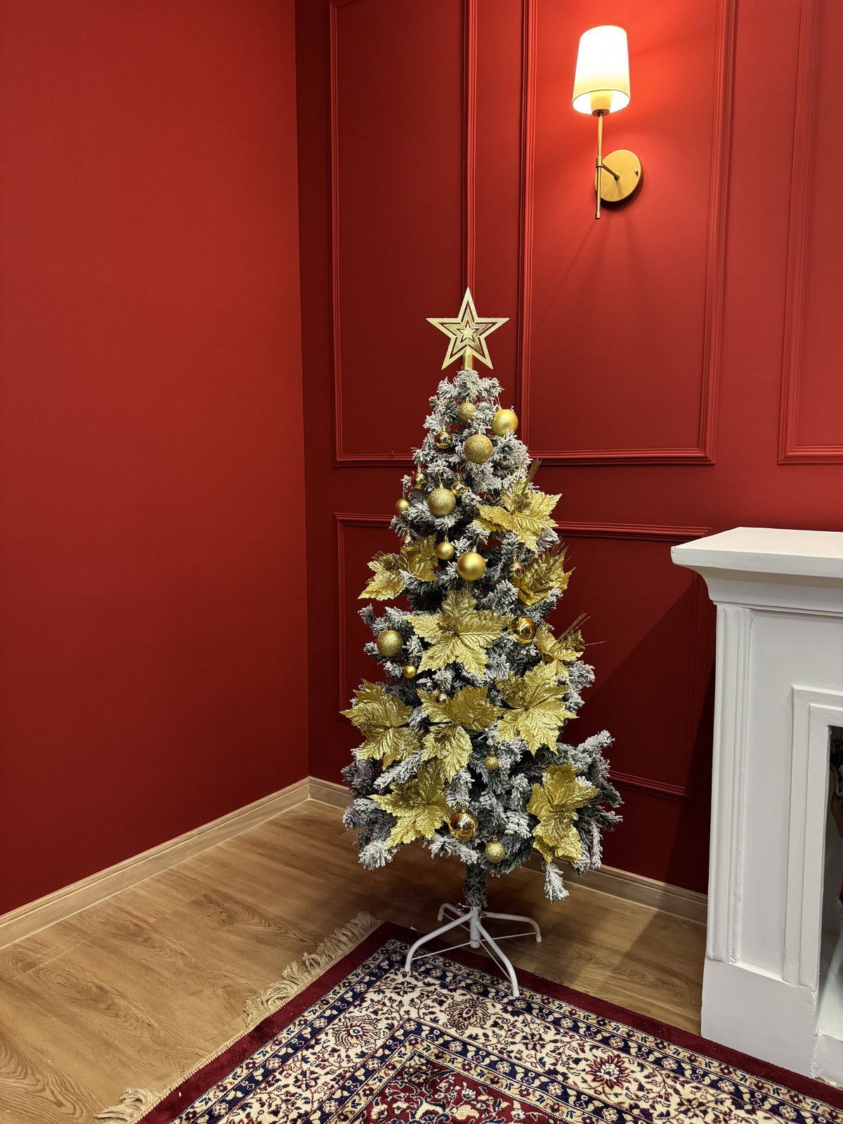 Árvore nevada de 1,50 m decorada com bolas e flores douradas e estrela no topo, em sala com parede vermelha e lareira branca