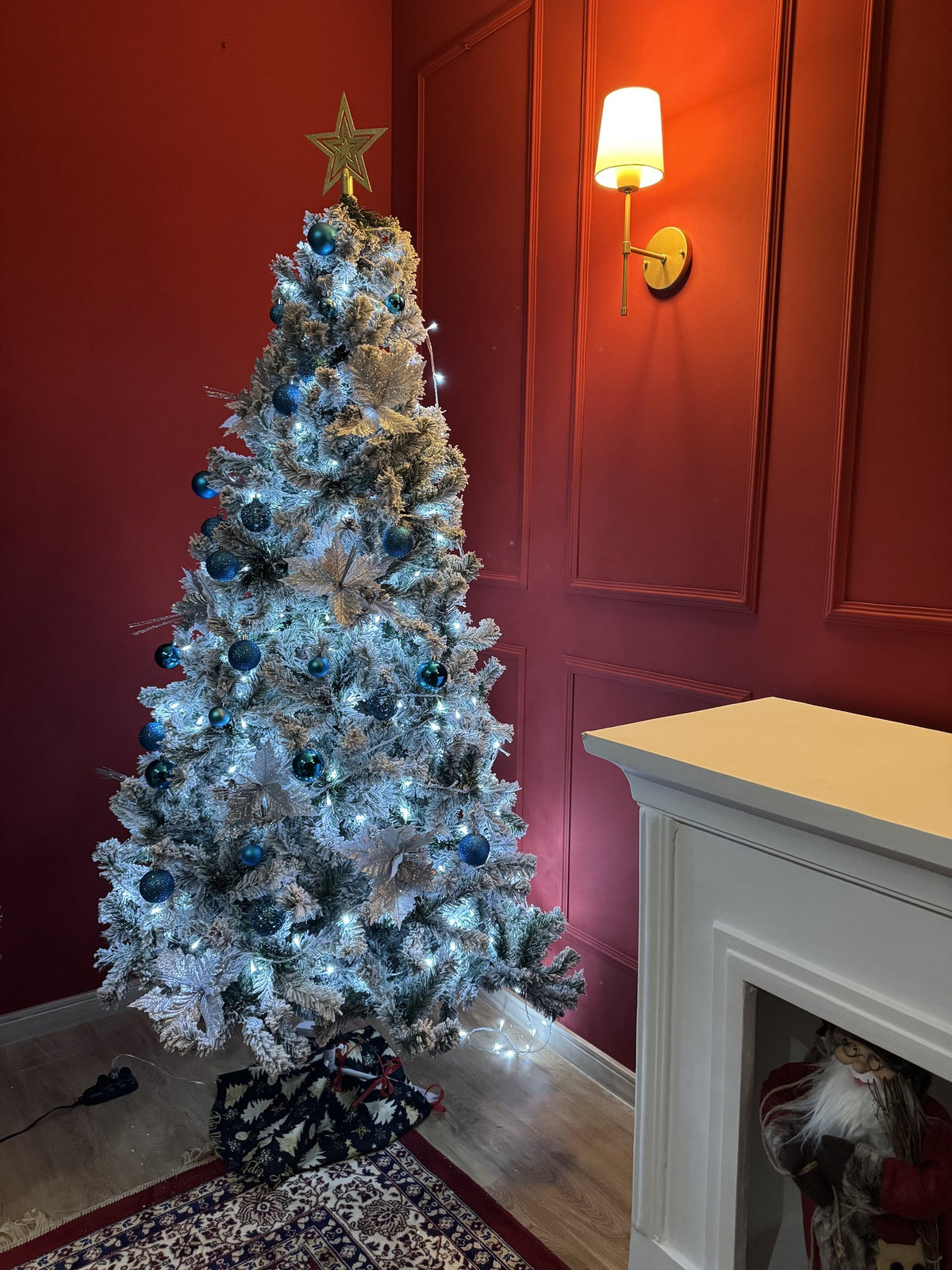 Árvore nevada de 1,80 m decorada com bolas azuis, laços dourados e luzes, ao lado de uma lareira branca