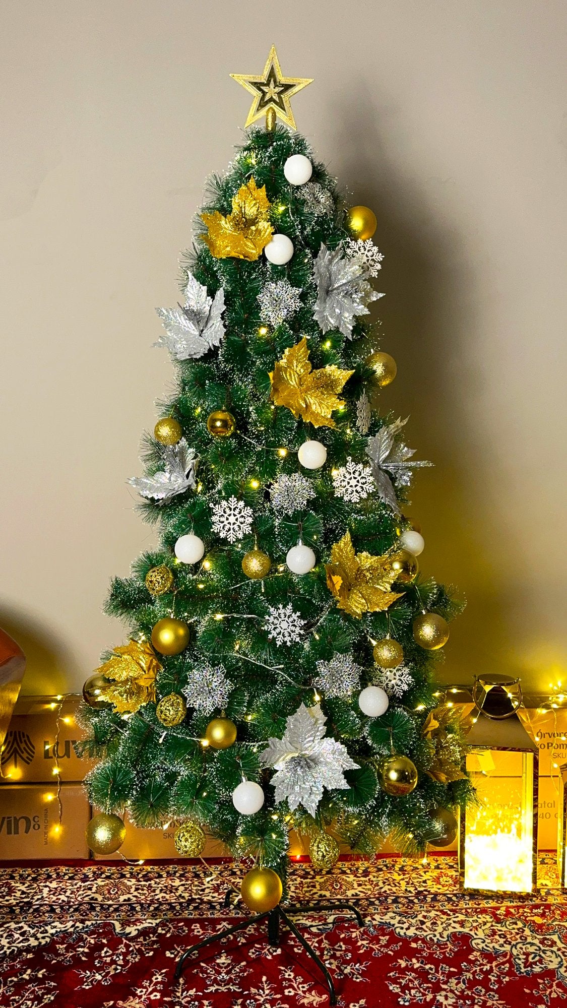 Árvore pompom com pontas nevadas de 1,80 m decorada com bolas douradas e prateadas, flores e estrela dourada, com lanterna e luzes ao fundo