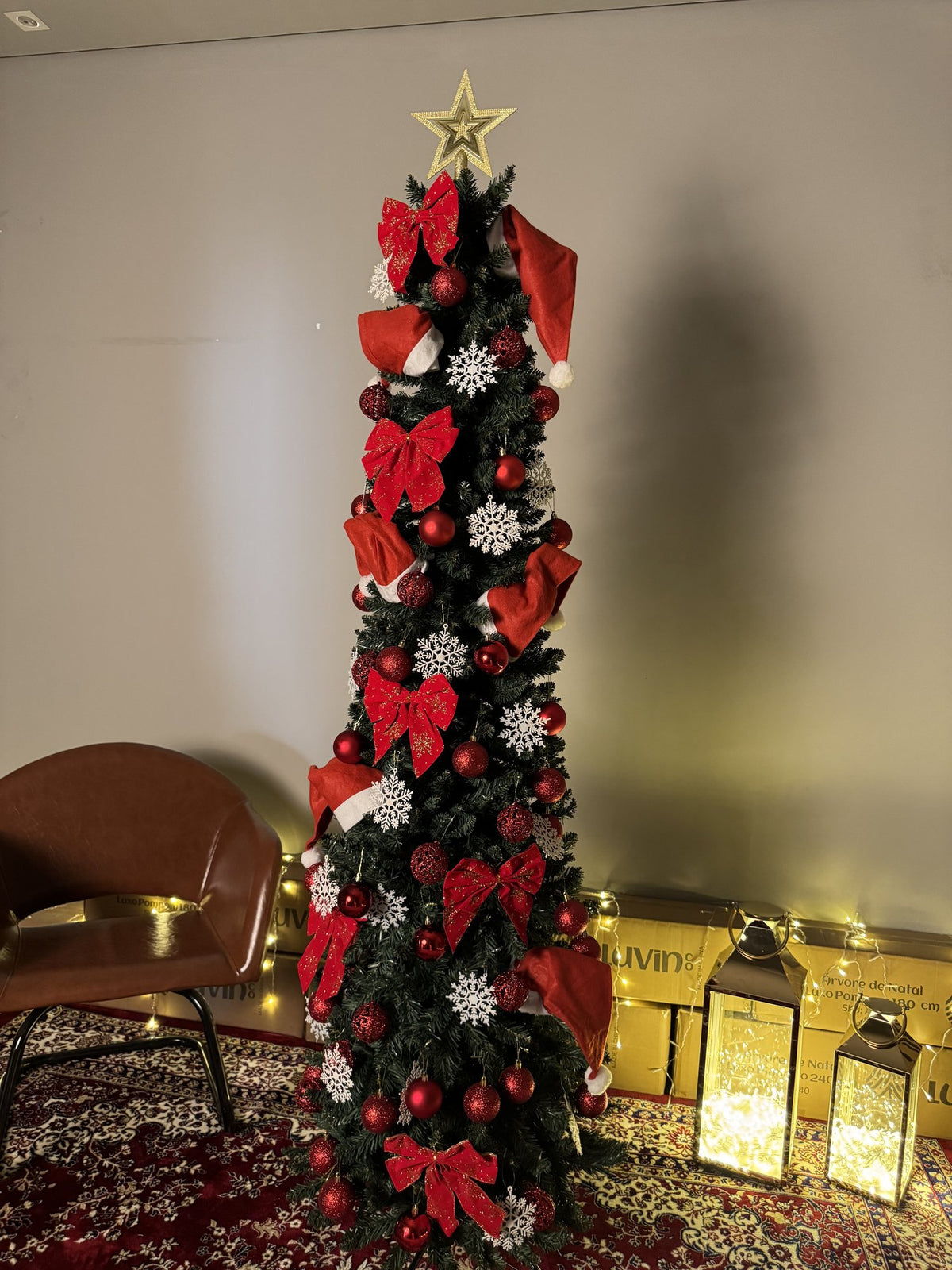 Árvore slim de 2,10 m decorada com laços vermelhos, bolas, flocos prateados e estrela dourada, ao lado de uma poltrona