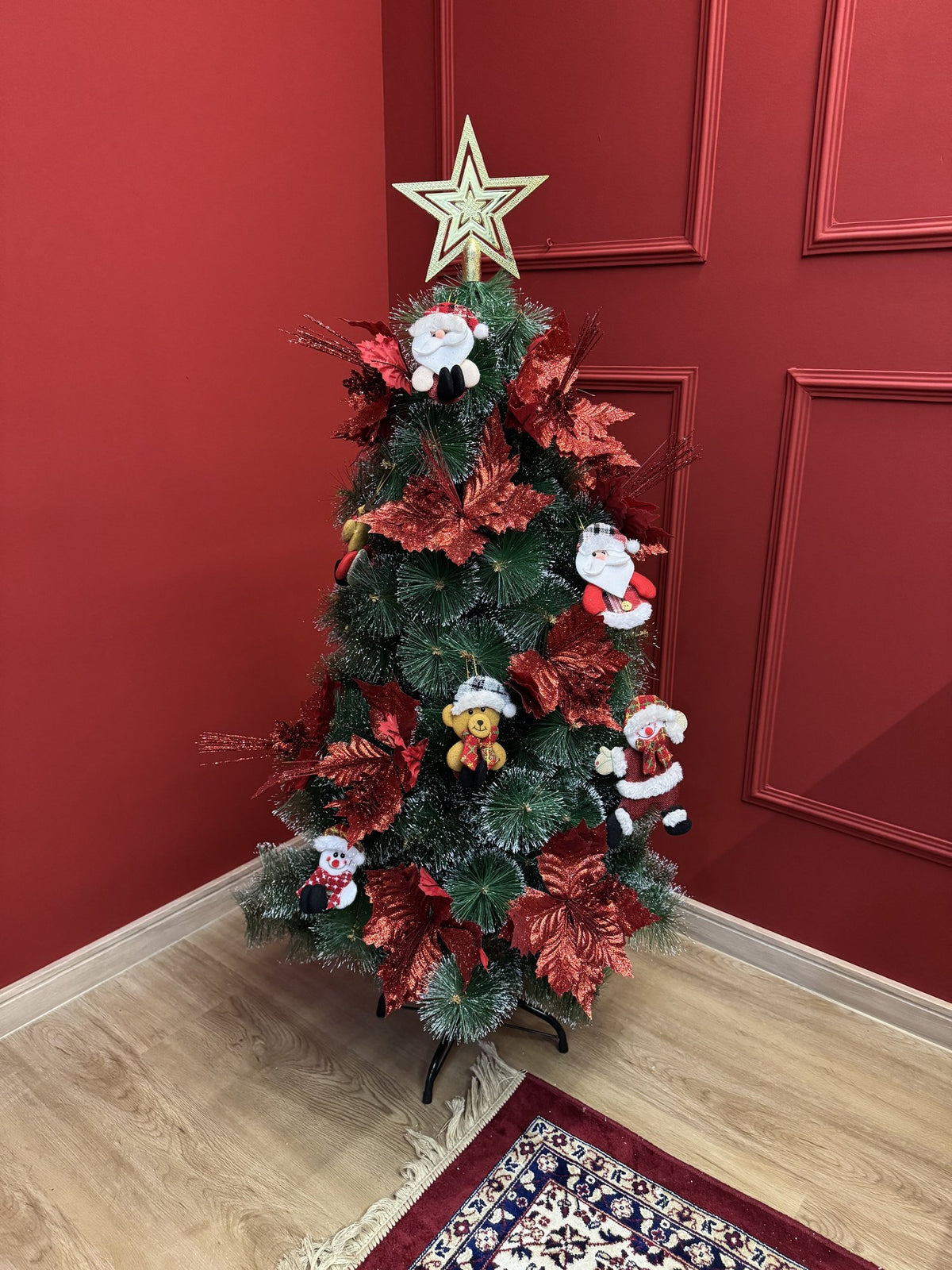 Árvore pompom com pontas nevadas de 1,20 m decorada com flores e laços vermelhos, bonecos de Papai Noel e estrela dourada, em canto de sala com parede vermelha