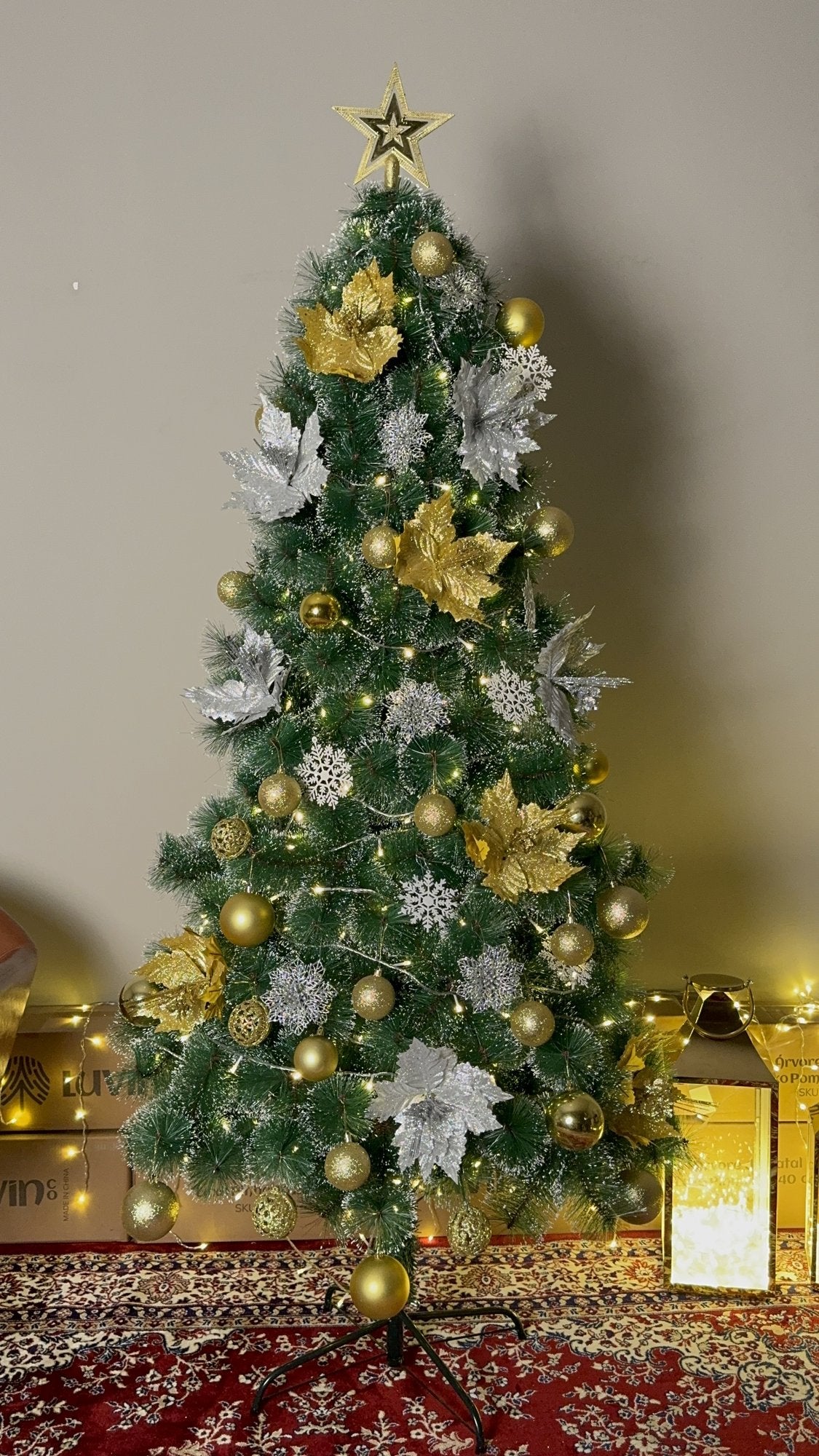 Árvore pompom com pontas nevadas de 2,10 m decorada com flores e bolas douradas e prateadas, com lanterna e luzes ao lado
