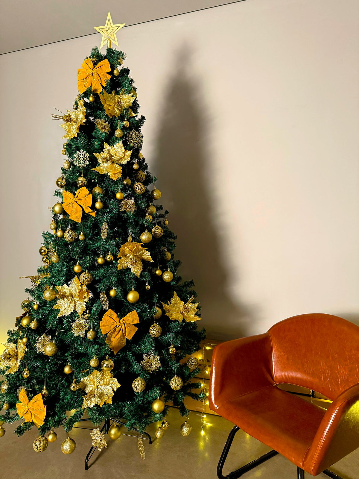 Pinheiro canadense tradicional de 2,40 m decorada com laços e flores amarelos, bolas douradas e estrela, ao lado de poltrona laranja