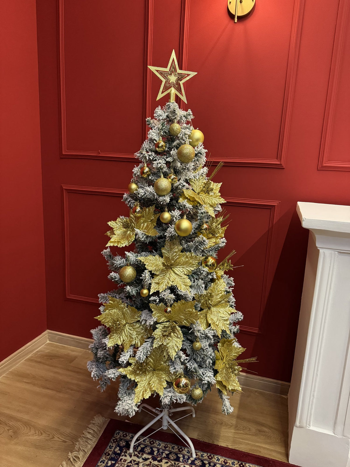 Árvore nevada de 1,50 m decorada com bolas e flores douradas e estrela no topo, vista de perto, em sala com parede vermelha