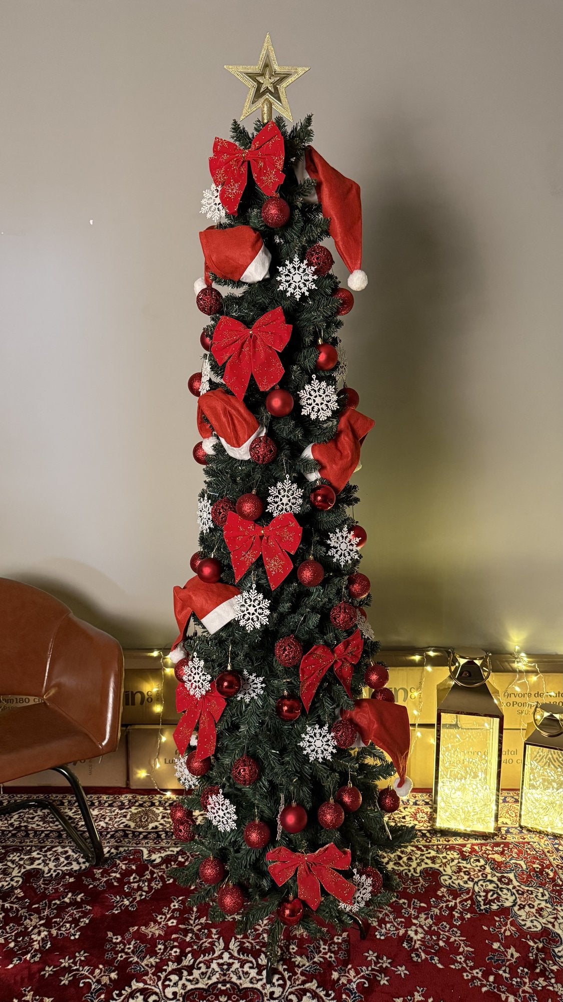 Árvore slim de 2,10 m decorada com laços vermelhos, bolas e flocos prateados, com lanternas e luzes ao fundo