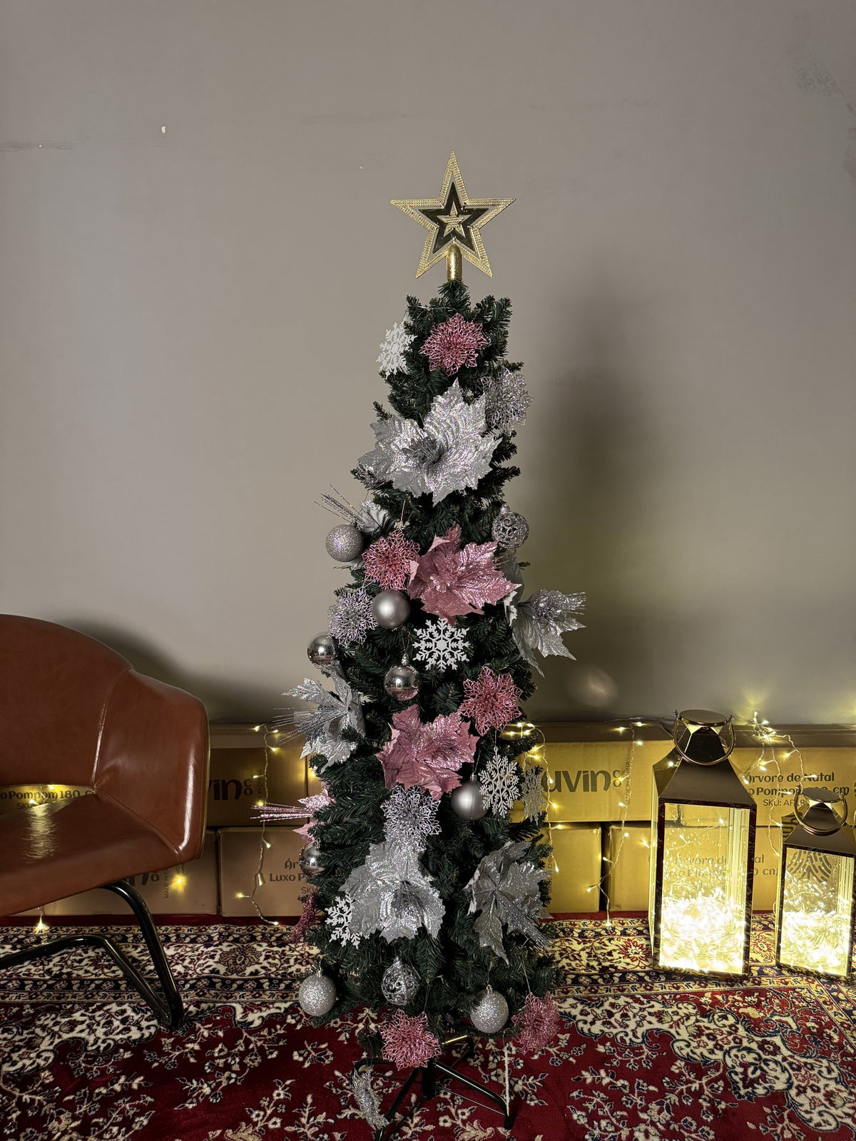 Árvore slim de 1,50 m decorada com flores rosa e prateadas e estrela dourada, em ambiente com poltrona e lanternas