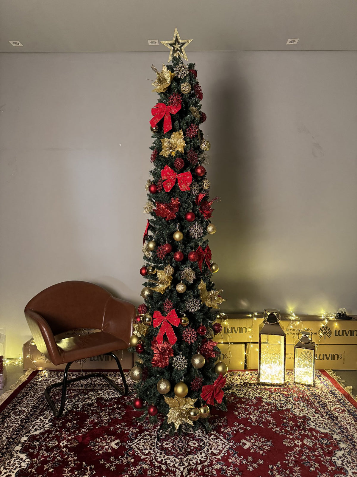Árvore slim de 2,40 m decorada com laços vermelhos, flores douradas e estrela, ao lado de uma poltrona de couro