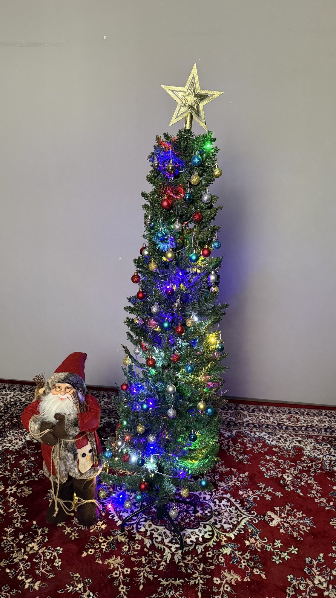 Árvore slim de 1,50 m decorada com pisca-pisca colorido, estrela dourada e Papai Noel ao lado