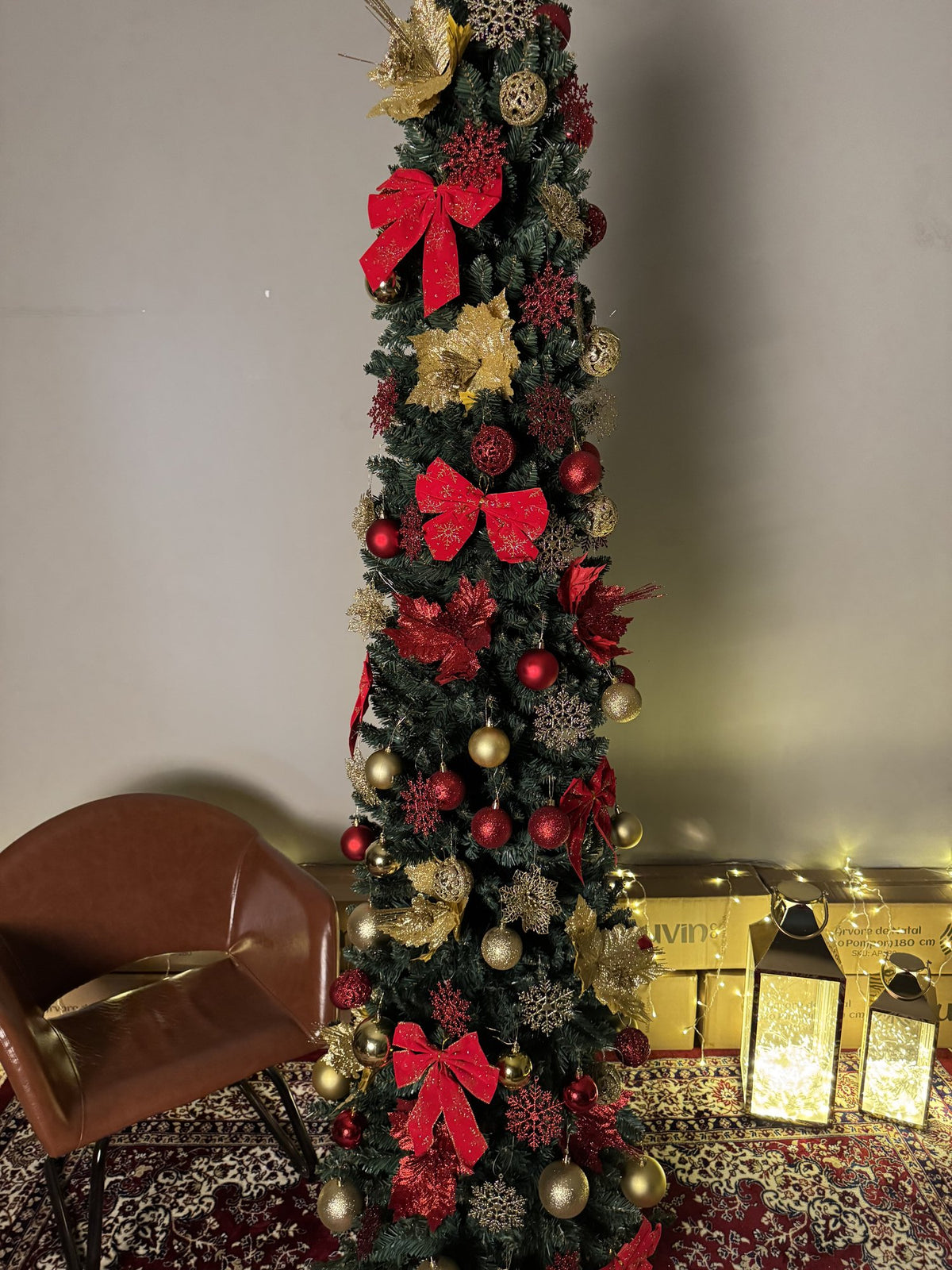 Árvore slim de 2,40 m decorada com laços vermelhos, flores e bolas douradas, ao lado de uma poltrona